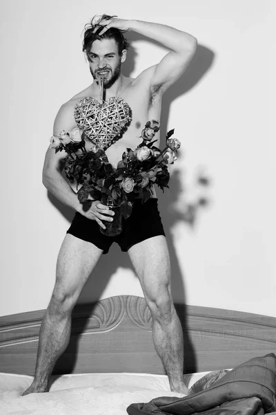 Guapo barbudo sexy macho hombre con elegante barba de pie en ropa interior en la cama cerca de manta roja sosteniendo rosas naranjas y decorativo corazón de San Valentín en teet — Foto de Stock