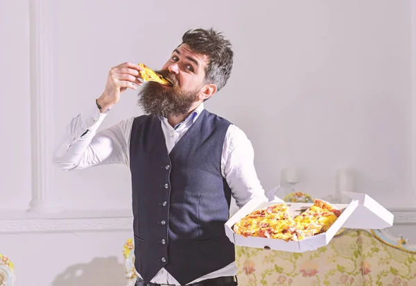 Macho in classic clothes hungry, on cheerful face, bites slice of pizza, eating, white background. Pizza delivery concept. Man with beard and mustache holds delivered box with tasty fresh hot pizza