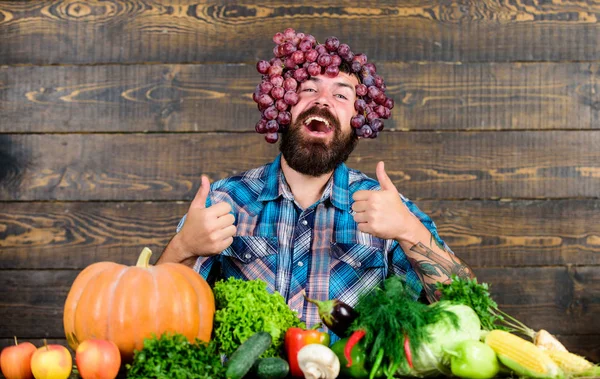 Farmer proud of grapes harvest. Fresh organic harvest. Grapes from own garden. Farming concept. Farmer bearded guy with homegrown harvest grapes put on head. Man hold grapes wooden background