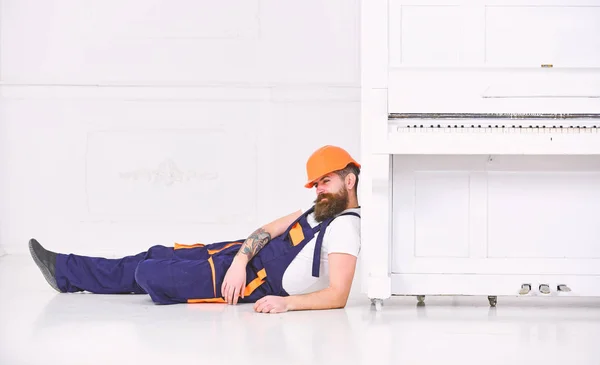 Loader lean on piano instrument. Man with beard, worker in overalls and helmet fall asleep tired, white background. Courier fall asleep while moving furniture, relocation. Tired worker concept