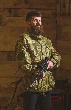 Sakallı kamuflaj elbiseler, ahşap iç arka plan giyer. Avcı, acımasız hipster silahla avlamaya hazır. Avcı kavramı. Maço gamekeepers evinde sıkı yüzünde