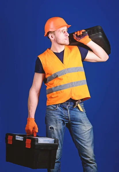 Homem de capacete, chapéu duro segura caixa de ferramentas e mala com ferramentas, fundo azul. Trabalhador, faz-tudo, reparador, construtor no rosto calmo carrega mala com ferramentas no ombro. Conceito de reparador equipado — Fotografia de Stock