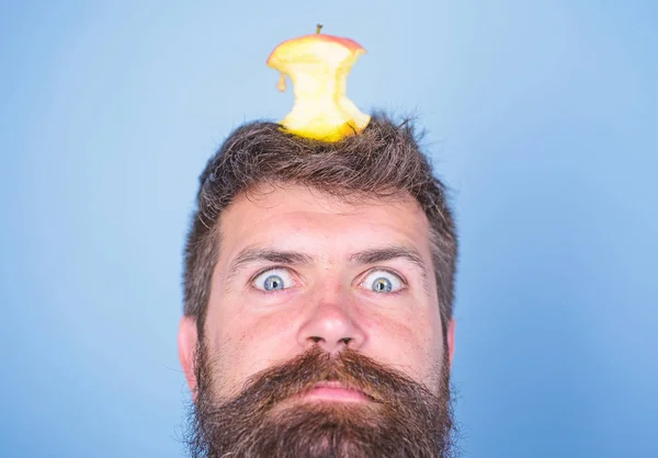 Hipster shocked face with apple stump target on head blue background, close up. Weight loss goal. Live target concept. Man handsome hipster long beard almost eaten apple stump on head as target