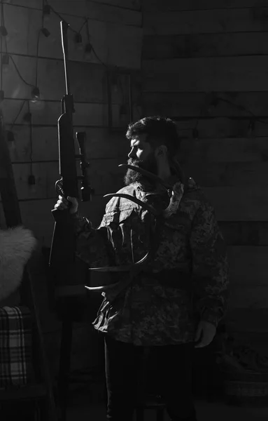 Concept Hunter. L'homme à la barbe porte des vêtements de camouflage, fond sombre. Macho sur le visage strict à la maison des gardiens prêts pour la chasse. Chasseur brutal avec pistolet et cornes de cerf, éclairé dans l'obscurité — Photo