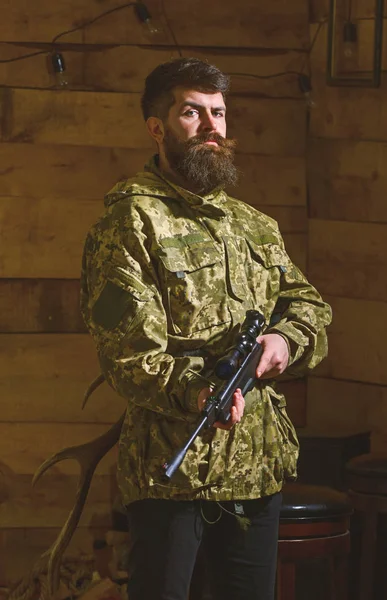 Hombre con barba lleva ropa de camuflaje, fondo interior de madera. Hunter, brutal hipster con arma lista para cazar. Concepto de cazador. Macho en la cara estricta en la casa de guardabosques —  Fotos de Stock