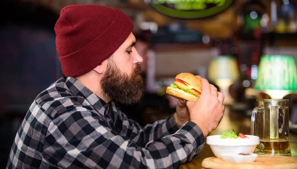 Hipster relaxing at pub. Pub is relaxing place to have drink and relax. Man with beard drink beer eat burger menu. Enjoy meal in pub. Brutal hipster bearded man sit at bar counter. High calorie snack