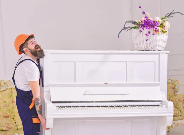 El mensajero entrega muebles, se muda, se traslada. Hombre con barba trabajador en casco y overol se levanta, esfuerzos para mover el piano, fondo blanco. El cargador mueve el instrumento de piano. concepto de cargas pesadas — Foto de Stock