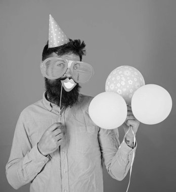 Hippi dev güneş gözlüğü kutluyor. Neşeli yüzündeki sakallı gülümseyen ağız sopa, kırmızı bir arka plan üzerinde tutar. Fotoğraf booth eğlence kavramı. Hava balonları ile parti şapkalı adamı kutluyor