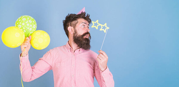 Man with beard on surprised face hold air balloons, light blue background. Hipster in star shaped glasses celebrates birthday. Guy in party hat with air balloons celebrates. Party attributes concept