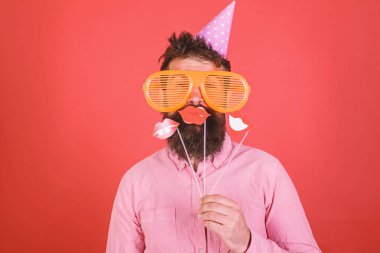 Parti şapkalı adamı kutlamak, fotoğraf sahne ile poz. Hippi dev güneş gözlüğü kutluyor. Duygusal çeşitlilik kavramı. Neşeli yüzündeki sakallı gülümseyen dudaklar sopa, kırmızı arka plan üzerinde tutar.