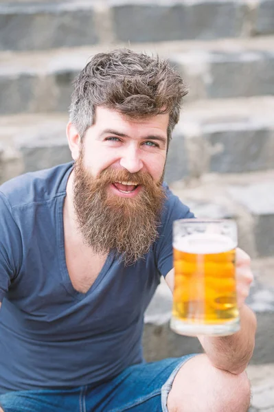 O hipster barbudo segura caneca de cerveja, bebe cerveja ao ar livre. Homem com barba e bigode na cara feliz, fundo pedregoso, desfocado. Conceito de cerveja artesanal. Hipster com barba longa parece relaxado — Fotografia de Stock