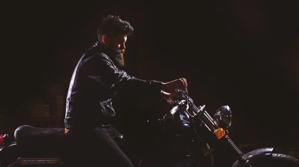 Night rider concept. Man with beard, biker in leather jacket sitting on motor bike in darkness, black background. Hipster, brutal biker in leather jacket riding motorcycle at night time, copy space — Stock Photo, Image