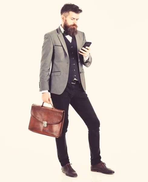 Brutal man ser upptagen. Skäggig man bär smart kostym och använder smartphone. Kille med allvarliga ansikte, fashionabla frisyr och portfölj. Business, internet, nya tachnology, framgång, manliga mode koncept — Stockfoto