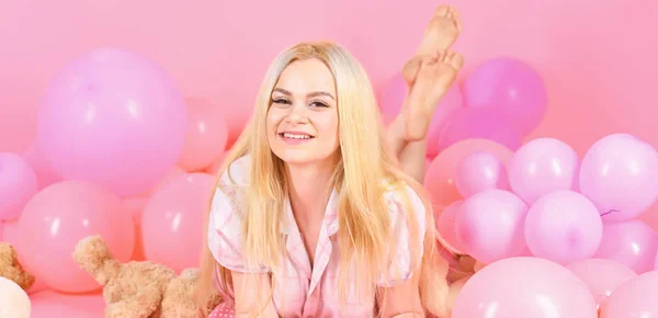 Girl in pajama, domestic clothes lay near air balloons, pink background. Birthday girl concept. Blonde on smiling face relaxing with teddy bear toy. Woman cute celebrate birthday with balloons