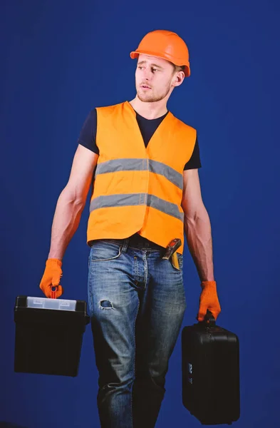 Homem de capacete, chapéu duro segura caixa de ferramentas e mala com ferramentas, fundo azul. Handyman, reparador na cara concentrada carrega sacos pesados com equipamento profissional. Conceito de reparador profissional — Fotografia de Stock