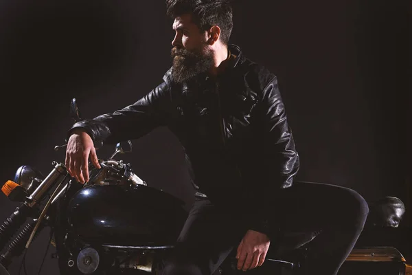 Brutality and masculine concept. Man with beard, biker in leather jacket lean on motor bike in darkness, black background. Macho, brutal biker in leather jacket stand near motorcycle at night time