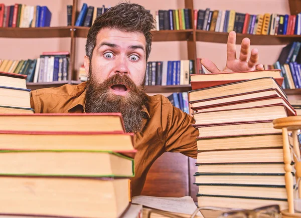 Mann mit schockiertem Gesicht zwischen Bücherstapeln, während er in der Bibliothek studiert, Bücherregale im Hintergrund. Lehrer oder Schüler mit Bart sitzen mit Brille am Tisch, defokussiert. Fristkonzept — Stockfoto