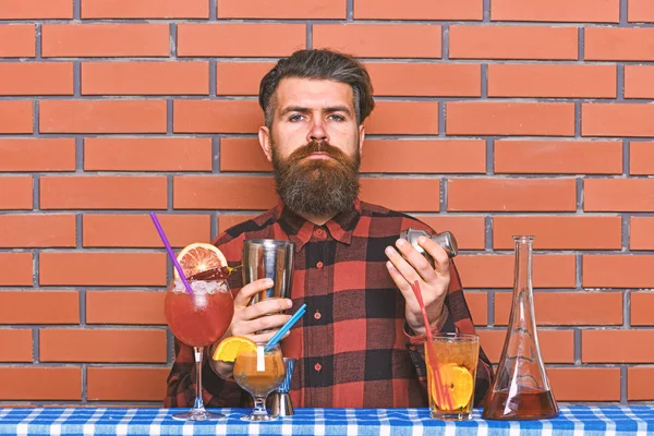 Alcoholic drinks concept. Barman with long beard and mustache and stylish hair on strict face holding shaker, makes alcoholic cocktail. Man in checkered shirt on brick wall background prepares drinks