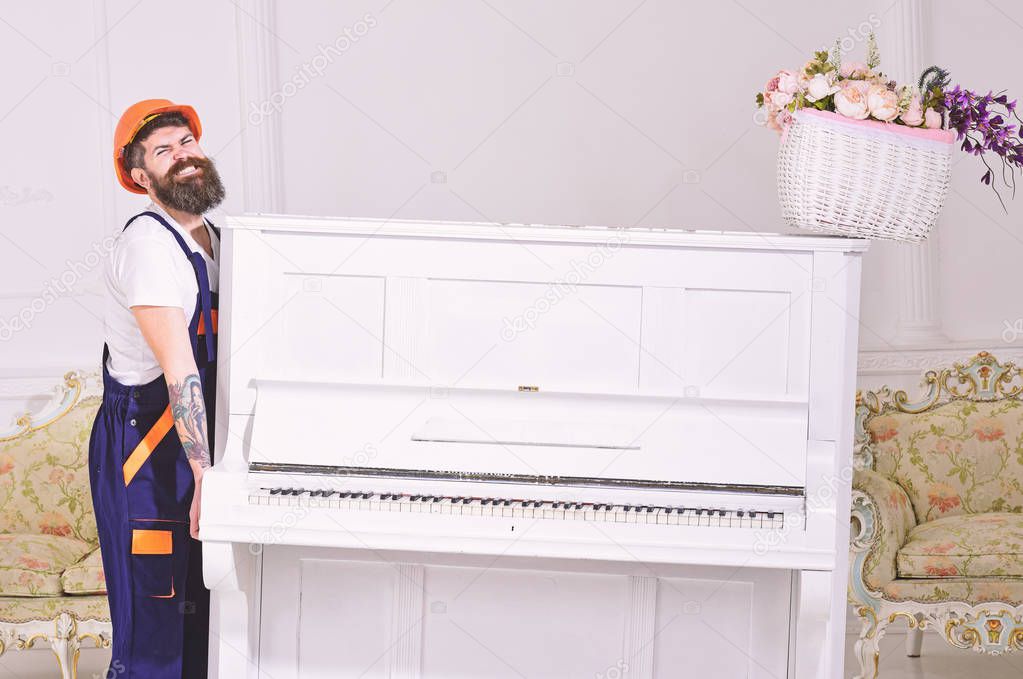 Loader moves piano instrument. Man with beard, worker in overalls and helmet lifts up piano, white background. Delivery service concept. Courier delivers furniture in case of move out, relocation