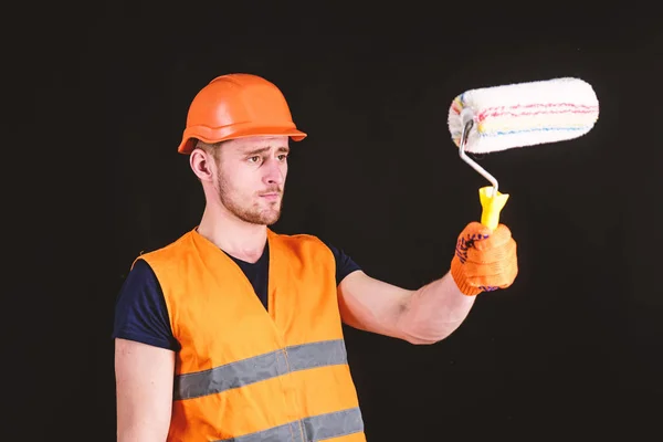 Decoratore pittura superficie di fronte a se stesso. Uomo in casco, cappello rigido tiene rullo di vernice, sfondo nero. Dipingere e rinnovare il concetto. Operaio, pittore, decoratore, appaltatore, costruttore sul volto occupato — Foto Stock