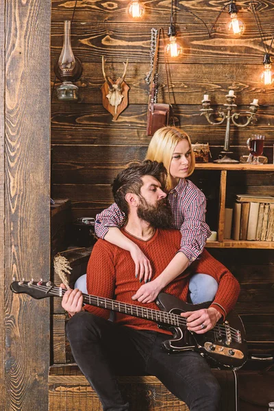 Couple in wooden vintage interior enjoy guitar music. Lady and man with beard on dreamy faces hugs and plays guitar. Couple in love spend romantic evening in warm atmosphere. Romantic evening concept