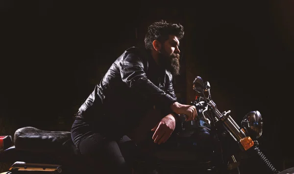 Masculinity concept. Macho, brutal biker in leather jacket riding motorcycle at night time, copy space. Man with beard, biker in leather jacket sitting on motor bike in darkness, black background