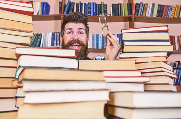 Homem no rosto feliz entre pilhas de livros na biblioteca, estantes de livros no fundo. Professor ou estudante com barba usa óculos, senta-se à mesa com livros, desfocado. Conceito de investigação científica — Fotografia de Stock