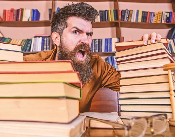 Учитель или ученик с бородой сидит за столом в очках, обезглавленный. Человек на кричащем лице между грудами книг, во время учебы в библиотеке, книжные полки на заднем плане. Концепция безумного ученого — стоковое фото