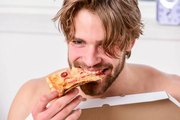 Guy holds pizza box sit bed in bedroom or hotel room. Hungry young man in underwear sits at home on a bed with a pizza box. Man bearded handsome bachelor eating cheesy food for breakfast in bed. — Stock Photo, Image