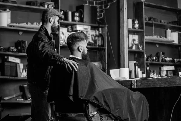 Hipster client got new haircut. Barber with bearded man looking at mirror, barbershop background. Haircut concept. Client and professional master checking result. Barber finished trimming