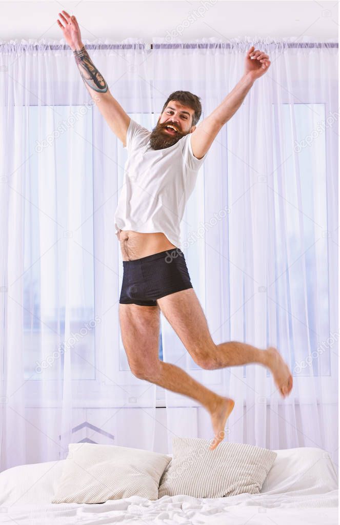 Man in shirt and underpants jumping on bed, white curtains on background. Guy on cheerful face full of energy in morning. Macho with beard jumps high in air. Full of strength and energy concept