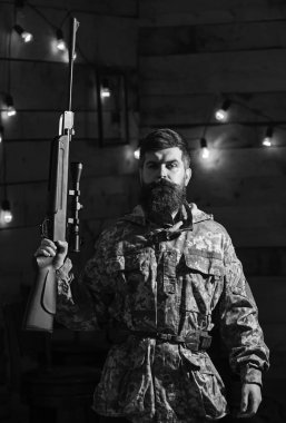 Hakem kavramı. Sakallı adam ahşap iç arka planda kamuflaj elbiseler giyer. Maço gamekeepers evinde sıkı yüzünde. Avcı, av için hazır elinde tabanca ile acımasız hipster