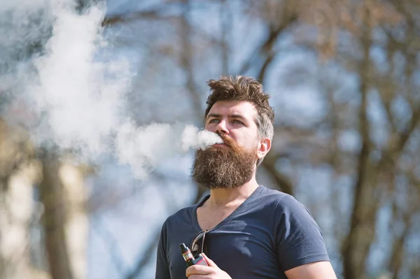Elektronikus cigaretta koncepció. Ember, hosszú szakállal és a felhők a füst néz ki nyugodt. Szakállas férfi dohányzik vape napsütéses napon. Férfi szakáll és bajusz nyugodt arc ágak háttere, defocused — Stock Fotó