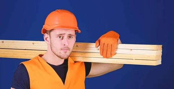 Tischler, Holzarbeiter, kräftiger Bauarbeiter mit nachdenklichem Gesicht trägt Holzbalken auf der Schulter. Mann mit Helm, Schirmmütze und Schutzhandschuhen hält Holzbalken, blauer Hintergrund. Holzwerkstoffkonzept — Stockfoto