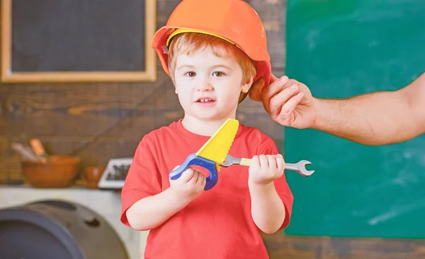 Little worker helping daddy in workshop. Kid playing with toy handsaw and spanner. Father holding protective helmet on sons head — Stock Photo, Image