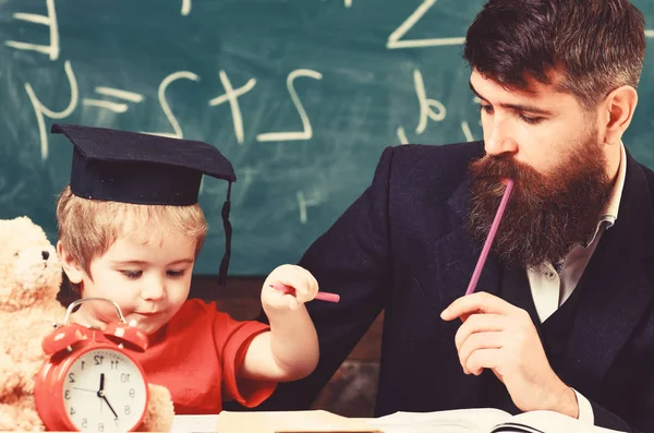 Father checking hometask, helps to boy, son. Hometask concept. Enthusiastic kid studying with teacher. Teacher in formal wear and pupil in mortarboard in classroom, chalkboard on background — Stockfoto