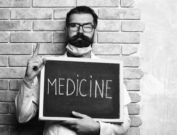 Bearded man, long beard. Brutal caucasian doctor or unshaven hipster, postgraduate student holding board with Medicine inscription in medical gown, glasses on beige brick wall studio backgroun