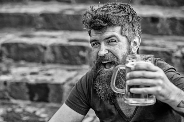 Hipster with long beard looks relaxed. Bearded hipster holds beer mug, drinks beer outdoor. Man with beard and mustache on happy face, stony background, defocused. Craft beer concept