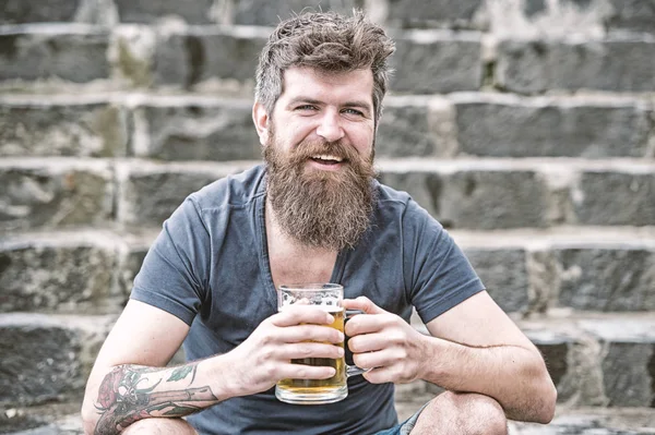 Cheerful man with beer in his hands sitting on stone staircase in downtown. Smiling young man with stylish beard relaxing after work. Friday night out, party mood