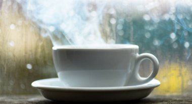 Kahve yağmurlu günde zevk. Taze yapılmış kahve beyaz fincan veya kupa pencere üzerinde. Yağmurlu bir gün kahve zamanında. Islak cam pencere ve sıcak kahve. Sonbahar bulutlu hava kafein içecek ile daha iyi