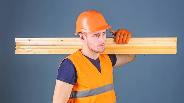 Carpintero, carpintero, fuerte constructor en la cara seria lleva la viga de madera en el hombro. Hombre con casco, sombrero duro y guantes de protección sostiene la viga de madera, fondo gris. Concepto de materiales de madera —  Fotos de Stock