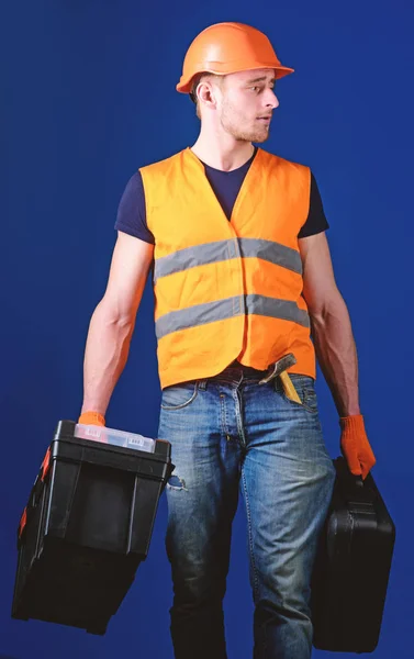 Professional repairman concept. Worker, handyman, repairman, builder on calm face carries bags with professional tools. Man in helmet, hard hat holds toolbox and suitcase with tools, blue background — Stock Photo, Image