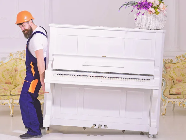 Loader moves piano instrument. Courier delivers furniture in case of move out, relocation. Man with beard, worker in overalls and helmet lifts up piano, white background. Delivery service concept