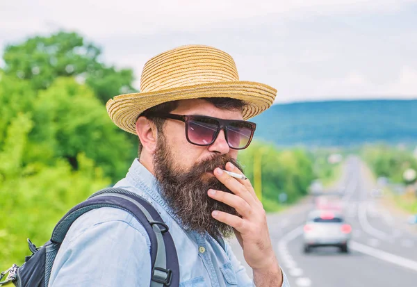 Cigarrillo antes de un largo viaje. El hombre con barba y bigote en sombrero de paja fumar cigarrillo, fondo de la carretera desenfocado. Fumar cigarrillo antes de viaje largo — Foto de Stock