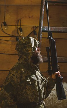 Avcı kavramı. Hunter, silahla acımasız hipster tüfek avcılık için hazırlayın. Maço gamekeepers evinde sıkı yüzünde. Sakallı adam başlıklı kamuflaj elbiseler, ahşap iç arka plan giyer
