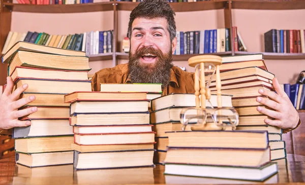 Homme sur le visage heureux entre des piles de livres, tout en étudiant à la bibliothèque, étagères de bibliothèque sur fond. Concept de bibliothécaire. Professeur ou élève barbu assis à table avec des livres, déconcentré — Photo