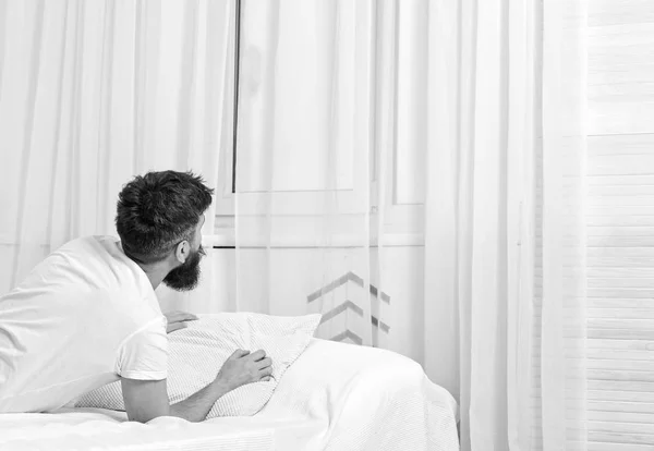 Hombre de camisa acostado en la cama despierto, cortina blanca en el fondo. Despierta y concepto matutino. Macho con barba y bigote relajante después de la siesta. El tipo se despierta por la mañana, mirando a la ventana —  Fotos de Stock