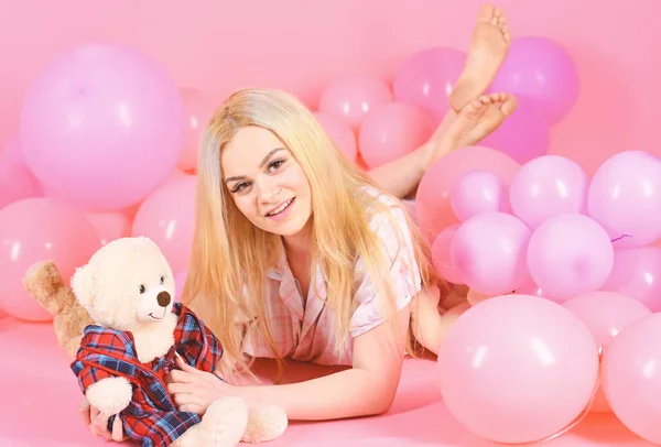Blonde on smiling face relaxing with teddy bear toy. Girl in pajama, domestic clothes lay near air balloons, pink background. Birthday girl concept. Woman cute celebrate birthday with balloons