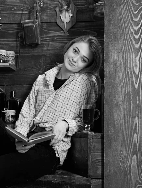 Conceito de relaxamento. Menina em roupa casual senta-se com livro no interior vintage de madeira. Estudante relaxante com livro e copo de vinho quente. Senhora no rosto sorridente em roupas xadrez parece casual — Fotografia de Stock
