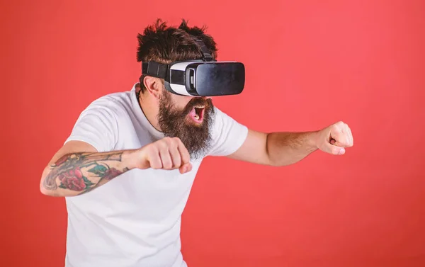 Hipster on excited face driving bike on high speed in virtual reality with modern digital gadget. Man with beard in VR glasses driving motorbike, red background. Virtual driving lessons concept